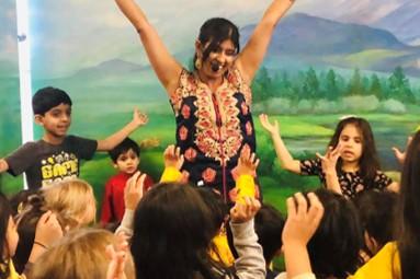 Indian presenter with arms held high surrounded by a group of children dancing.